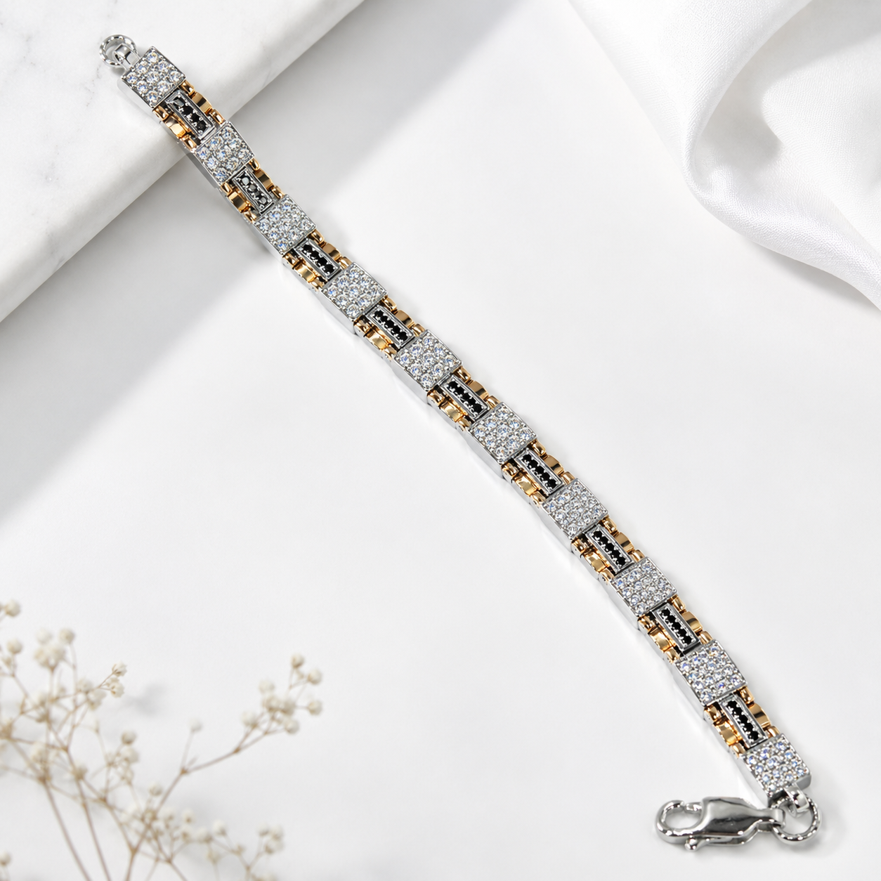 A silver-toned link bracelet with alternating rows of clear pavé stones and small gold and black accents, displayed diagonally on a white marble and satin background with dried flowers.