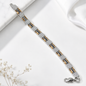 A silver-toned link bracelet with alternating rows of clear pavé stones and small gold and black accents, displayed diagonally on a white marble and satin background with dried flowers.