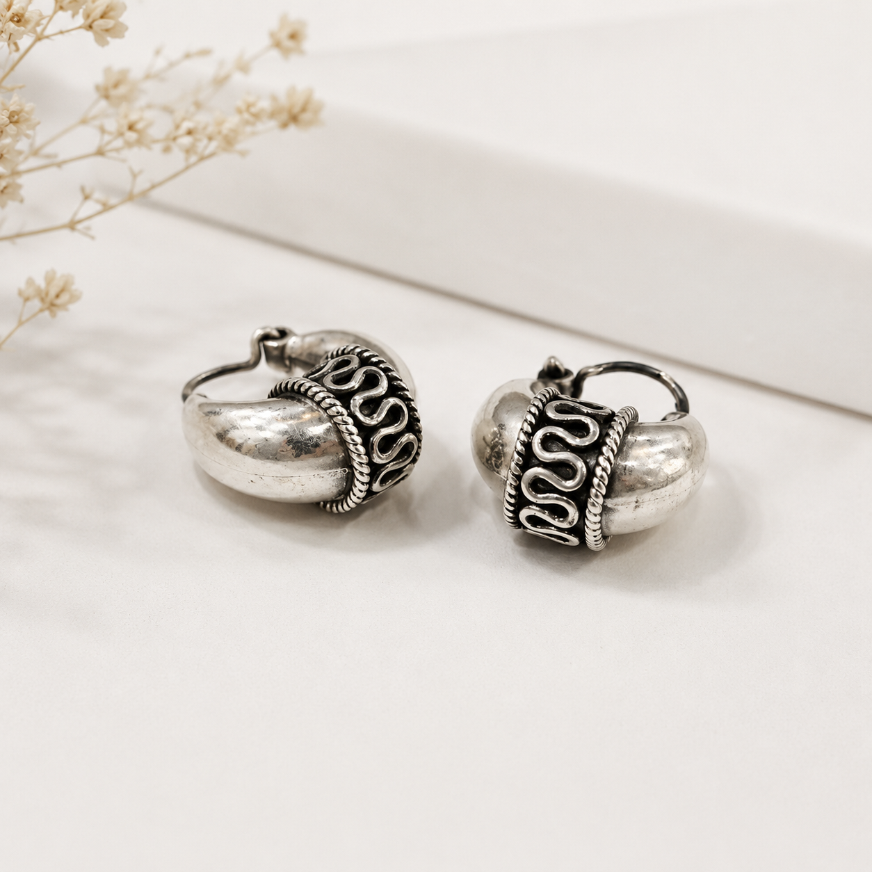 A pair of silver-toned huggie hoop earrings with textured metallic bodies and dark oxidized scrollwork details, displayed on a clean white surface with dried flowers in the background.