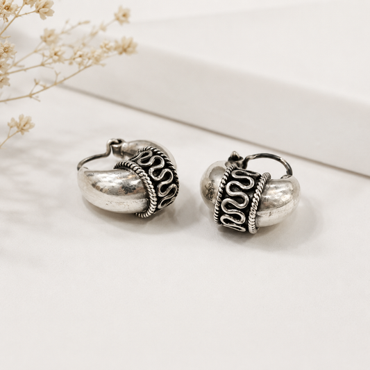 A pair of silver-toned huggie hoop earrings with textured metallic bodies and dark oxidized scrollwork details, displayed on a clean white surface with dried flowers in the background.