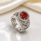 A silver-toned ring featuring a large oval red gemstone surrounded by a halo of small clear stones, with additional channel-set clear stones along the band, photographed on a soft white surface with a draped cream fabric background.