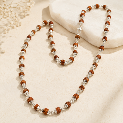 A beaded necklace with brown rudraksha beads and silver-toned textured spacer beads, arranged on a light beige surface with a white stone prop.